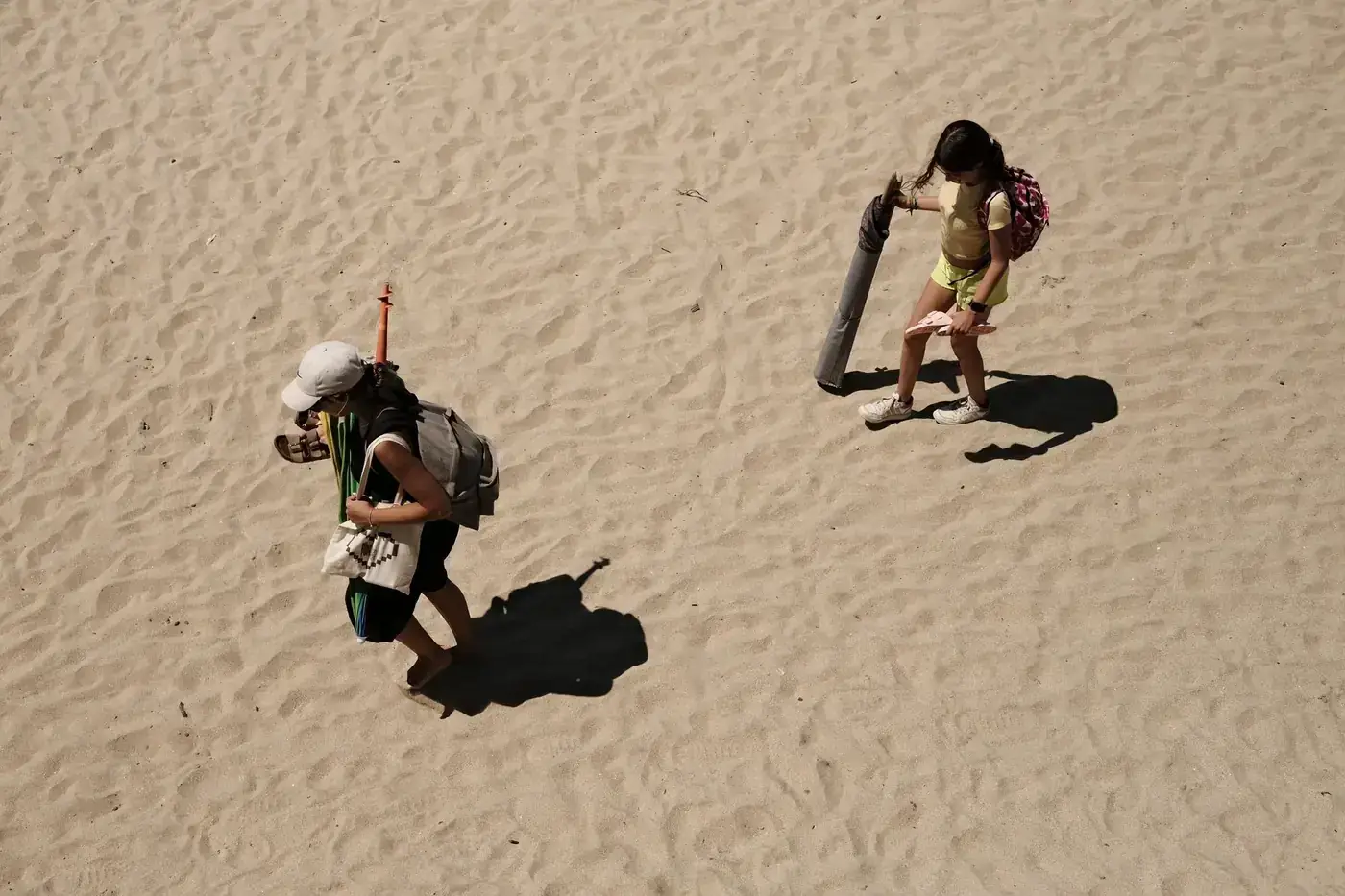Two people carrying bags walk across a sandy beach under sunlight, creating long shadows.