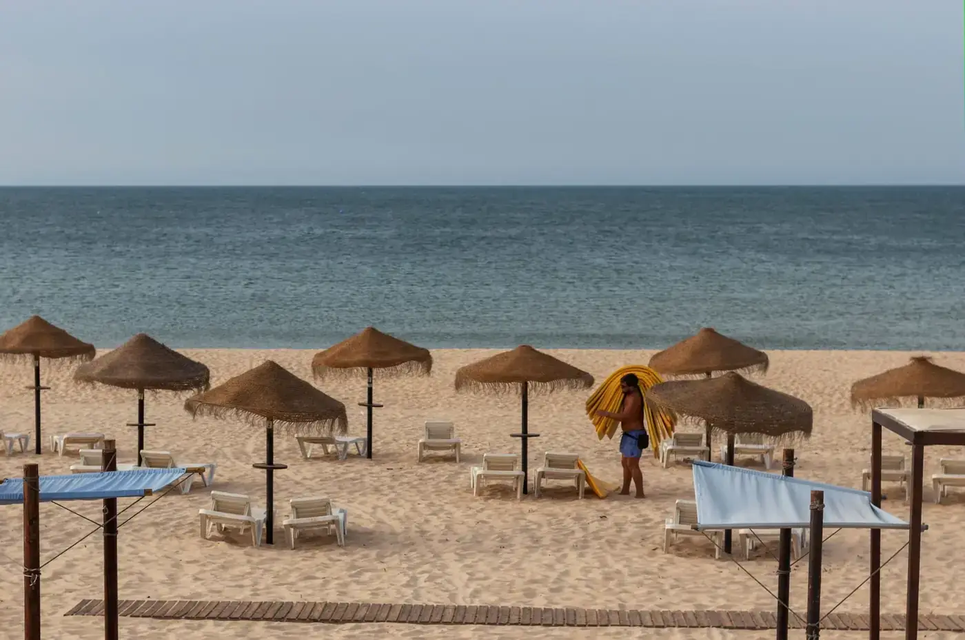 A tranquil beach scene with straw umbrellas and a person preparing sun loungers by the sea.