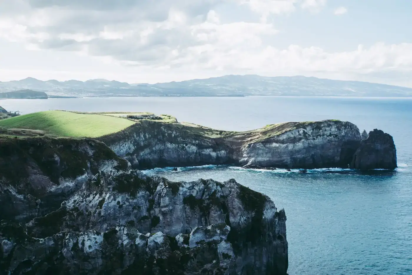 Majestic coastline with dramatic cliffs and vibrant green fields meeting azure blue ocean waters under a cloudy sky.