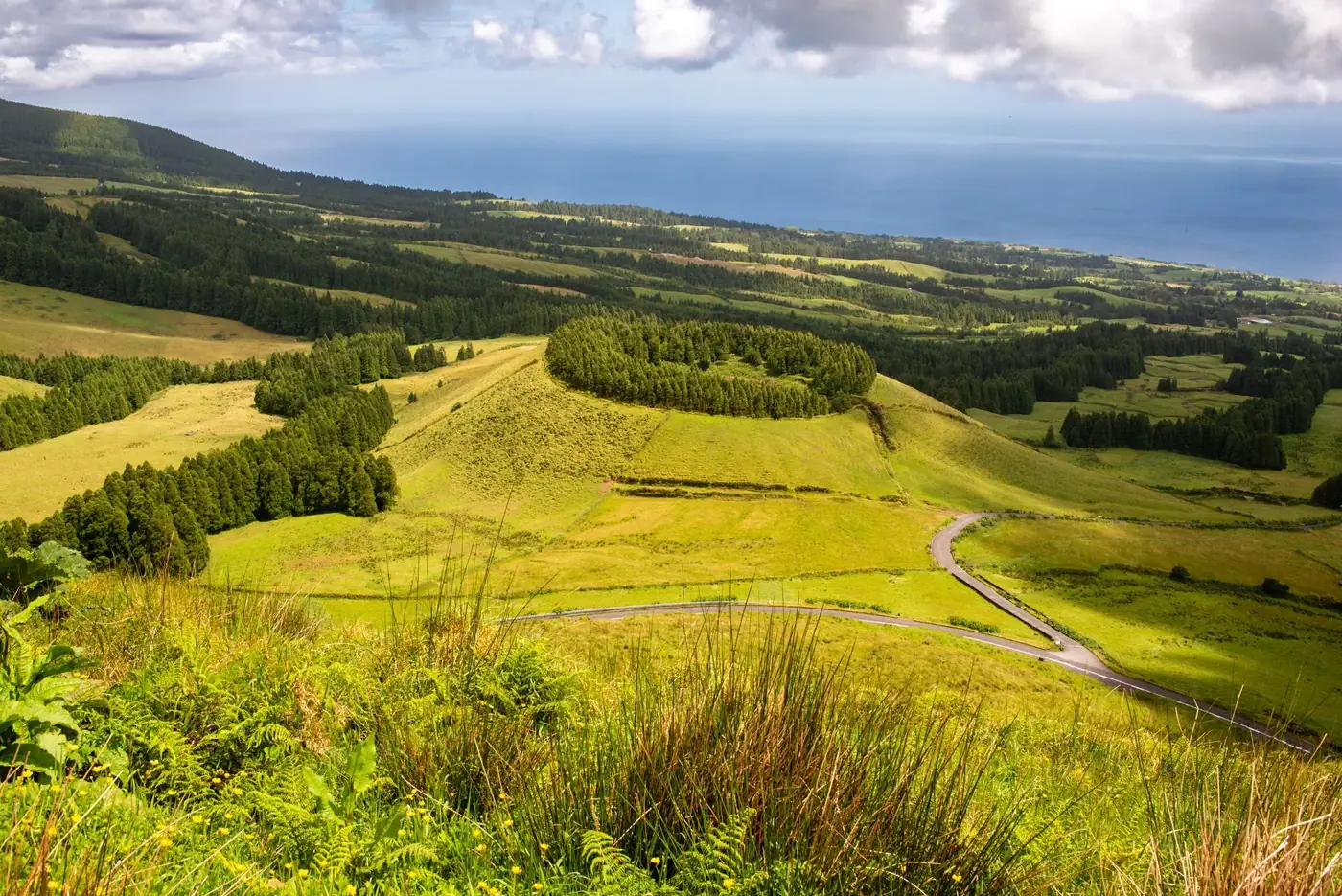 Lush green landscape of Azores with rolling hills and distant ocean view under cloudy sky.