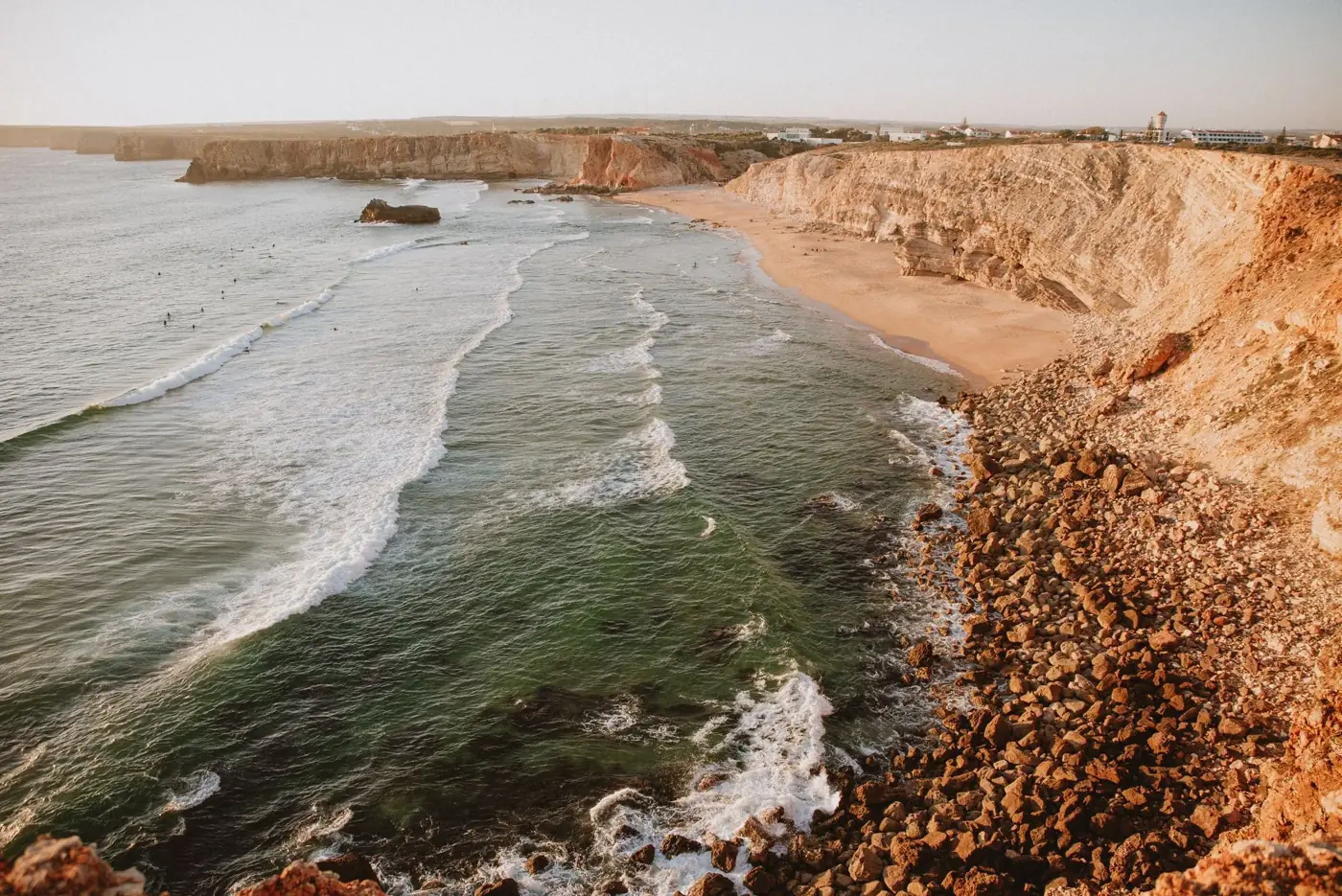 Aerial view of a rugged cliff seacoast with waves crashing onto the rocks and beach at sunset.